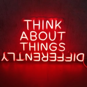 Könnte beinhalten: Ein rotes Neon-Schild zeigt die Worte "THINK ABOUT THINGS DIFFERENTLY" in einer fetten, serifenlosen Schriftart. Das Schild leuchtet vor einem dunklen Hintergrund und erzeugt einen auffälligen visuellen Kontrast. Die Neonröhren sind hell und gleichmäßig beleuchtet.