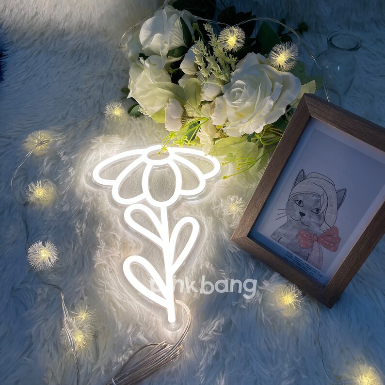 May include: White neon sign shaped like a daisy flower with a stem and leaves. The sign is lit up and is sitting on a white fluffy surface. There is a framed picture of a cat with a bow tie in the background.
