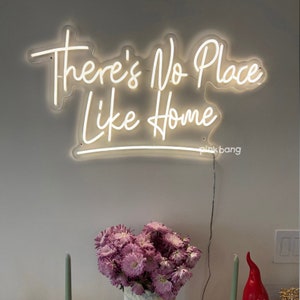 May include: White neon sign with the text "There's No Place Like Home" on a white wall. The sign is lit up and there are flowers in a vase below it.