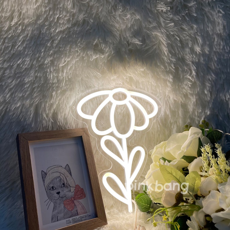 May include: White neon sign shaped like a daisy flower with a stem and leaves. The sign is lit up and is sitting on a white fuzzy surface.