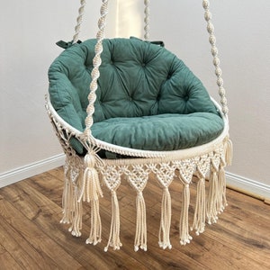 May include: A white macrame hanging chair with a green cushion. The chair has a fringe border and tassels.