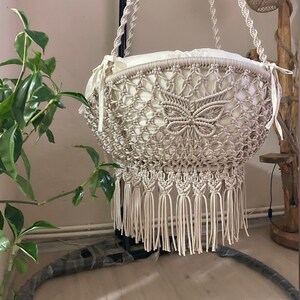 Op de afbeelding: Een beige macramé hangstoel met een wit kussen. De stoel heeft ingewikkelde knoopmotieven en een gefranjerde onderkant. Hij hangt aan een zwart metalen frame, wat een bohemien uitstraling creëert.