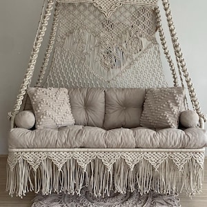 May include: A beige macrame swing with a fringed base and canopy. The swing features a cushioned seat with two throw pillows and a matching round rug. The intricate woven design adds a bohemian touch to the indoor space.
