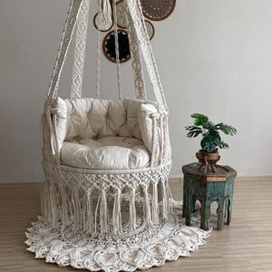 Peut inclure: Un fauteuil suspendu en macramé crème avec un coussin touffeté et des franges. La chaise est suspendue à un crochet en métal. Un tapis en macramé assorti est en dessous. Une petite table d'appoint turquoise vieillie supporte une plante en pot.