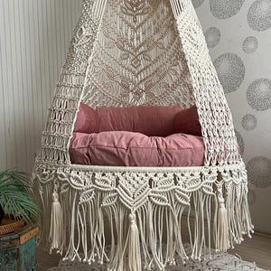 以下が含まれることがあります： クリーム色のマクラメハンギングチェア。ピンクのクッションと枕が付いています。椅子はフリンジの底があり、織られたキャノピーから吊り下げられています。お揃いの丸いラグが下にあります。