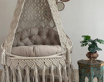 May include: A beige macrame hanging chair with a tufted cushion, fringed edges, and a canopy. The chair is suspended from the ceiling. A small, distressed turquoise side table holds a potted plant. A matching rug is beneath the chair.
