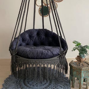 Peut inclure: Un fauteuil suspendu en macramé gris foncé avec un coussin touffeté et des franges. La chaise est suspendue au plafond et repose sur un tapis rond en macramé gris assorti. Des décorations murales sont en arrière-plan.
