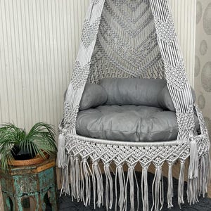 Peut inclure: Un fauteuil suspendu en macramé gris avec un coussin rond et deux coussins. La chaise a un bas à franges et est suspendue à un anneau métallique. Un tapis gris est sous la chaise.