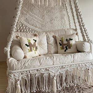 Peut inclure: Un fauteuil suspendu en macramé blanc avec une jupe et un auvent à franges. Le siège comporte deux coussins décoratifs avec des lettres brodées et un tapis de couleur neutre en dessous. La chaise est suspendue à une corde.