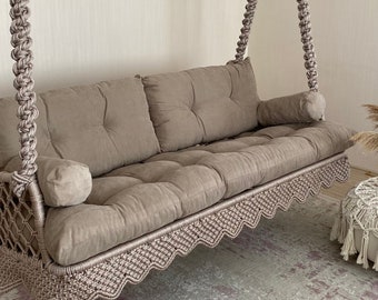 Könnte beinhalten: Ein beiges Makramee-Schaukelsofa mit einer gesteppten Sitzfläche und Rückenlehne sowie zwei Kissen. Die Schaukel ist mit dicken, geflochtenen Seilen an der Decke aufgehängt. Das Sofa hat einen dekorativen, wellenförmigen Rand.