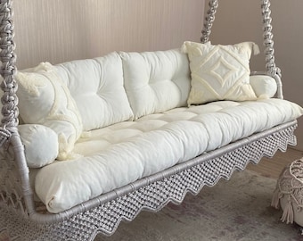 Könnte beinhalten: Ein graues Makramee-Hängesofa mit weißen Kissen und einem weißen Dekokissen. Die Schaukel hängt an einer weißen Decke und steht vor einer weißen Wand mit vertikalen Linien.