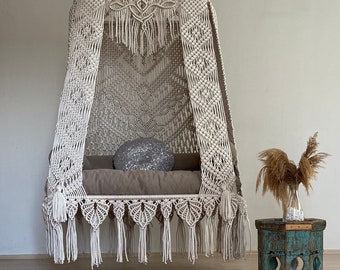 May include: A white macrame hanging chair with a fringe border and a pillow. The chair is hanging from the ceiling and is in a room with a wooden floor.