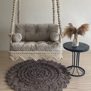 May include: A beige macrame swing chair with a gray velvet cushion and two pillows. The swing chair is hanging from the ceiling and is over a gray crocheted rug.