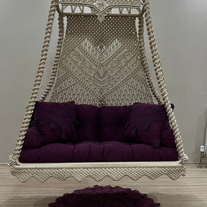 Op de afbeelding: Een beige macramé hangstoel met een paars kussen en twee kussens. De stoel hangt aan het plafond en is gesuspendeerd boven een paars gehaakt kleed.