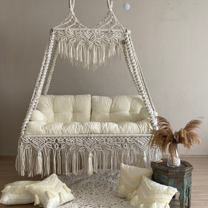 May include: A white macrame hanging chair with a white cushion and fringe. The chair is hanging from the ceiling with a chain. There are three white throw pillows on the floor in front of the chair.
