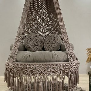 May include: A beige macrame hanging chair with a thick cushion and two round pillows. The chair is hanging over a beige crocheted rug.