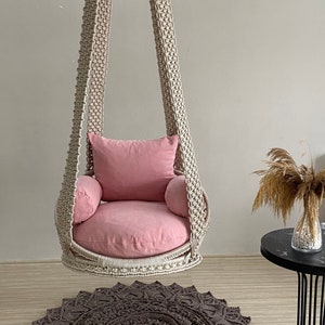 May include: A macrame hanging chair with a pink cushion and a brown crocheted rug underneath.