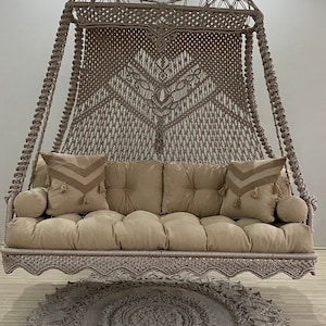 May include: A macrame swing with a beige cushion and pillows, suspended from the ceiling. The swing is made of woven rope and features a decorative fringe along the bottom. A matching round rug sits beneath the swing.