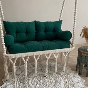 May include: A white macrame swing chair with green cushions and tassels. The swing chair is hanging from the ceiling and is in front of a white wall. A white crocheted rug is on the floor in front of the swing chair.