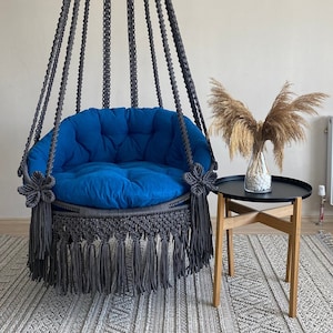 May include: A gray macrame hanging chair with a blue cushion and fringe. The chair is hanging from the ceiling and is in front of a white wall. A small wooden table with a black top is in the foreground.