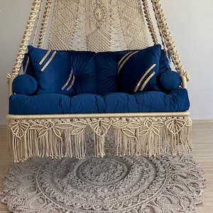 Op de afbeelding: Een hangstoel van macramé met een blauw fluwelen kussen en twee kussens. De stoel hangt aan een haak aan het plafond en is omgeven door een beige macramé tapijt.