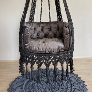 May include: A black macrame hanging chair with a dark gray cushion and fringe. The chair is hanging from the ceiling and is sitting on a gray macrame rug.