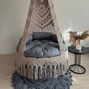 May include: A beige macrame hanging chair with a grey cushion and fringe. The chair is hanging from the ceiling in a room with a light wood floor.