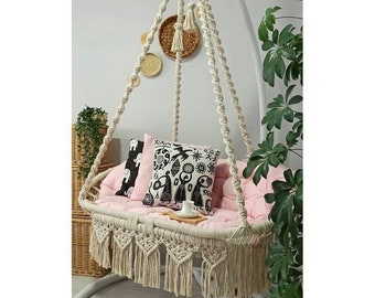 May include: A white macrame hanging chair with a pink cushion and two black and white patterned pillows. The chair has a fringe trim and is hanging from a white metal stand.