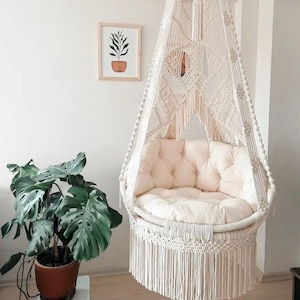 Op de afbeelding: Een witte macramé hangstoel met een wit kussen. De stoel hangt aan het plafond en heeft een franje rond de onderkant.