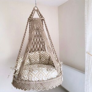 Pu&ograve; includere: Una sedia a dondolo in macram&egrave; beige con un cuscino a motivi bianchi e beige. La sedia &egrave; sospesa dal soffitto e ha un bordo a frangia.