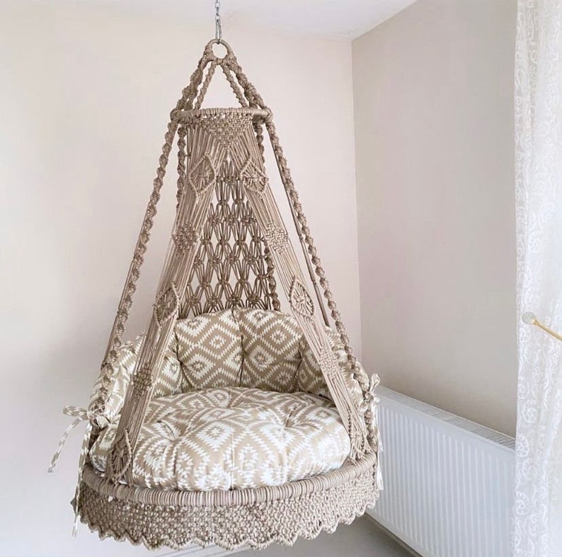Pu&ograve; includere: Una sedia a dondolo in macram&egrave; beige con un cuscino a motivi bianchi e beige. La sedia &egrave; sospesa dal soffitto e ha un bordo a frangia.