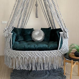 May include: A gray macrame hanging chair with a green velvet cushion and a silver sequin pillow. The chair has a fringe border and is hanging from a ceiling hook.