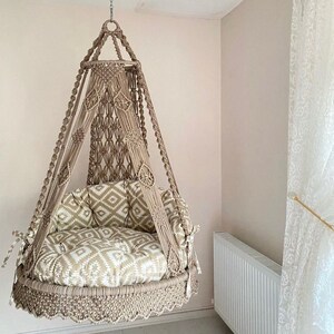 Pu&ograve; includere: Una sedia a dondolo in macram&egrave; beige con un cuscino a motivi. La sedia &egrave; sospesa dal soffitto e ha un bordo a frangia.