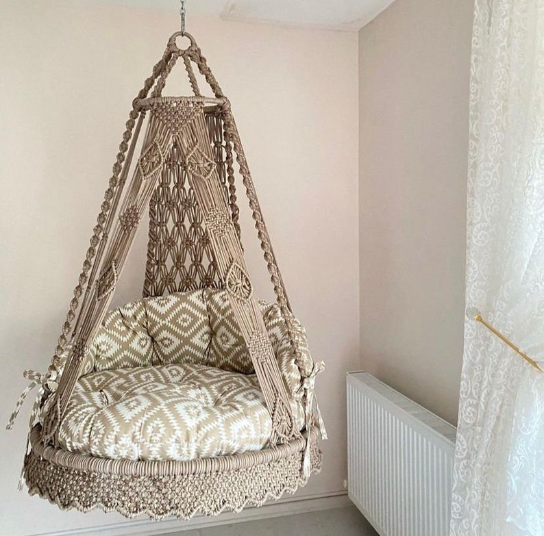 Pu&ograve; includere: Una sedia a dondolo in macram&egrave; beige con un cuscino a motivi. La sedia &egrave; sospesa dal soffitto e ha un bordo a frangia.