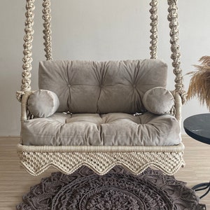 May include: A white macrame swing chair with a gray velvet cushion and two pillows. The swing chair is hanging from the ceiling and is over a gray and white crocheted rug.
