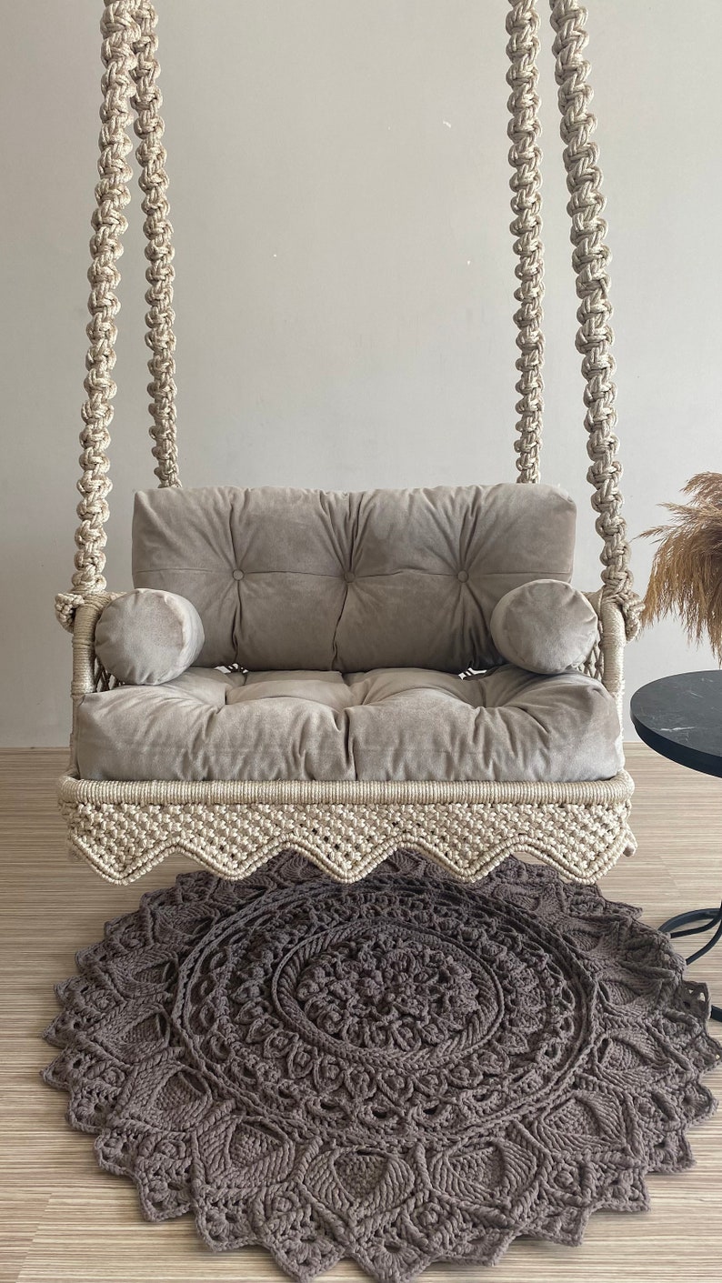 May include: A white macrame swing chair with a gray velvet cushion and two pillows. The swing chair is hanging from the ceiling and is over a gray and white crocheted rug.