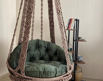 Könnte beinhalten: Ein brauner Makramee-Hängesessel mit einem getufteten grünen Kissen. Der Stuhl ist von der Decke abgehängt. Im Hintergrund ist ein kleines Bücherregal mit Büchern und Dekorationsgegenständen zu sehen. Ein schönes Wohnaccessoire.