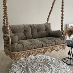 May include: A brown rope swing with a beige cushion and a white crocheted rug underneath. The swing is hanging from the ceiling and has a scalloped edge.