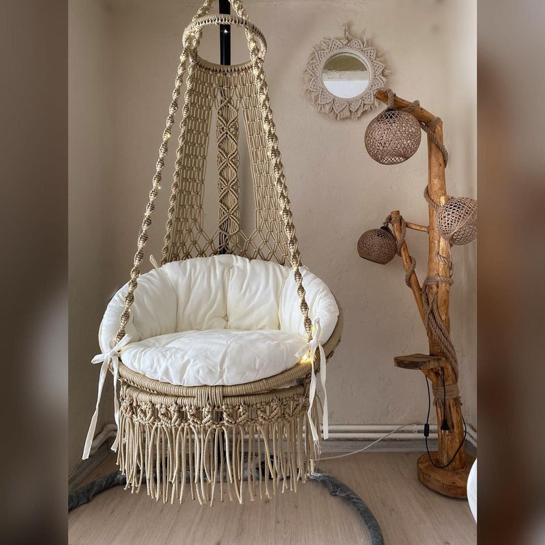 May include: A beige macrame hanging chair with a white cushion and fringe detail. The chair is hanging from a black metal stand. The chair is in a room with a wooden floor and a wooden lamp stand with a beige shade.