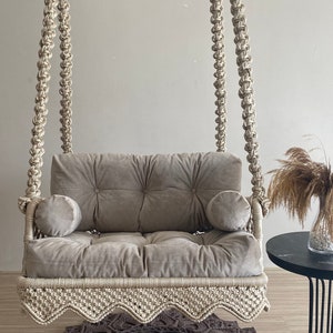 May include: A beige macrame swing chair with a gray velvet cushion and two pillows. The swing chair is hanging from the ceiling and is in front of a gray wall. A brown crocheted rug is on the floor in front of the swing chair.