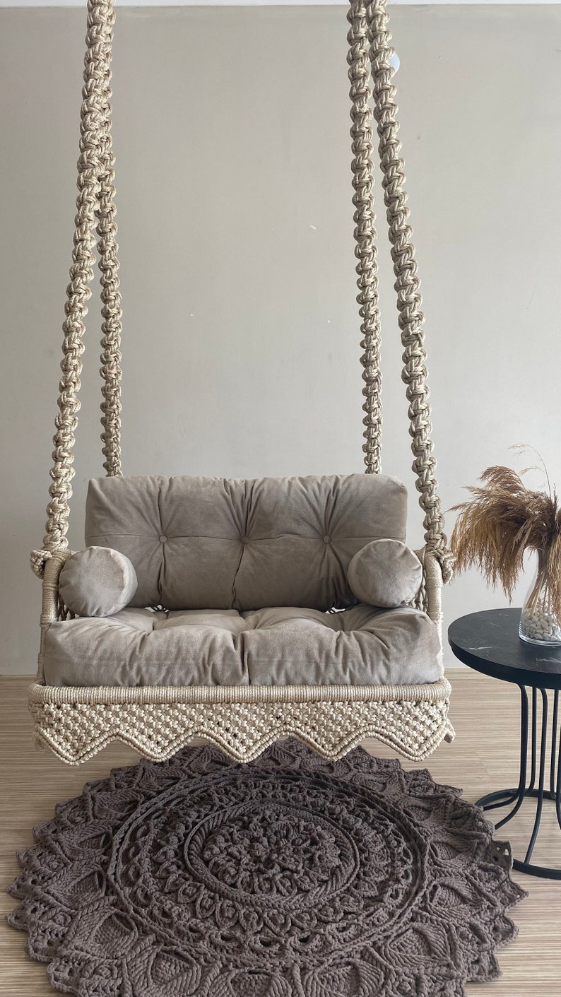 May include: A beige macrame swing chair with a gray velvet cushion and two pillows. The swing chair is hanging from the ceiling and is in front of a gray wall. A brown crocheted rug is on the floor in front of the swing chair.