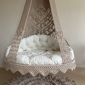 May include: A beige macrame hanging chair with a white cushion and a matching beige macrame rug on the floor.