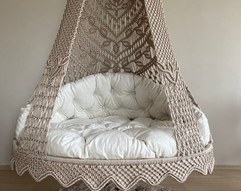 May include: A beige macrame hanging chair with a white cushion and a matching beige macrame rug on the floor.