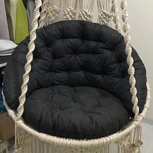 May include: A white macrame hanging chair with a black padded cushion. The chair is made of thick rope and has a fringe around the bottom.