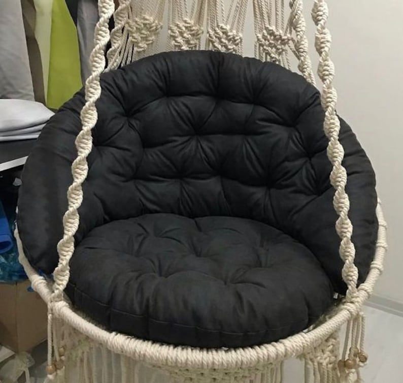 May include: A white macrame hanging chair with a black padded cushion. The chair is made of thick rope and has a fringe around the bottom.