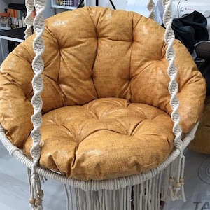 May include: A white hanging chair with a thick, round, mustard yellow cushion. The chair is made of woven rope and has a fringe around the bottom.