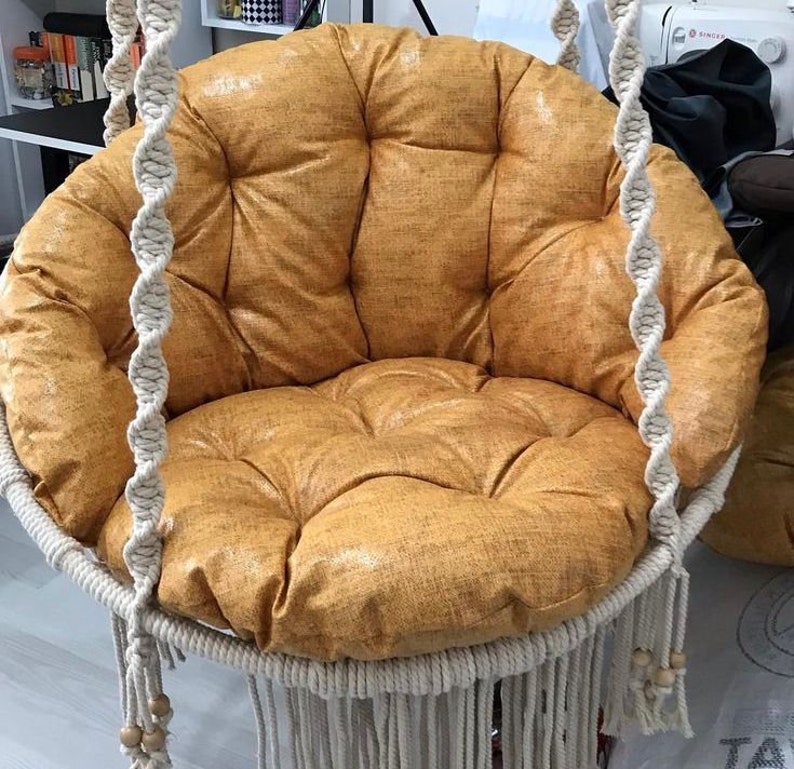 May include: A white hanging chair with a thick, round, mustard yellow cushion. The chair is made of woven rope and has a fringe around the bottom.