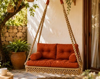 May include: A hanging bench swing featuring a rectangular seat with thick, tufted rust-orange cushions and matching cylindrical side bolsters. The base is wrapped in natural tan rope with a decorative macrame fringe, suspended by thick braided ropes.