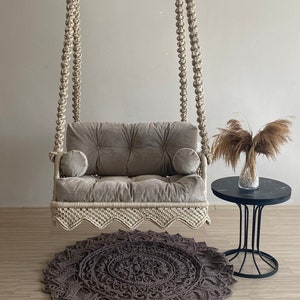 May include: A macrame swing chair with a gray cushion and two pillows. The swing is hanging from the ceiling and is over a brown rug with a circular pattern.