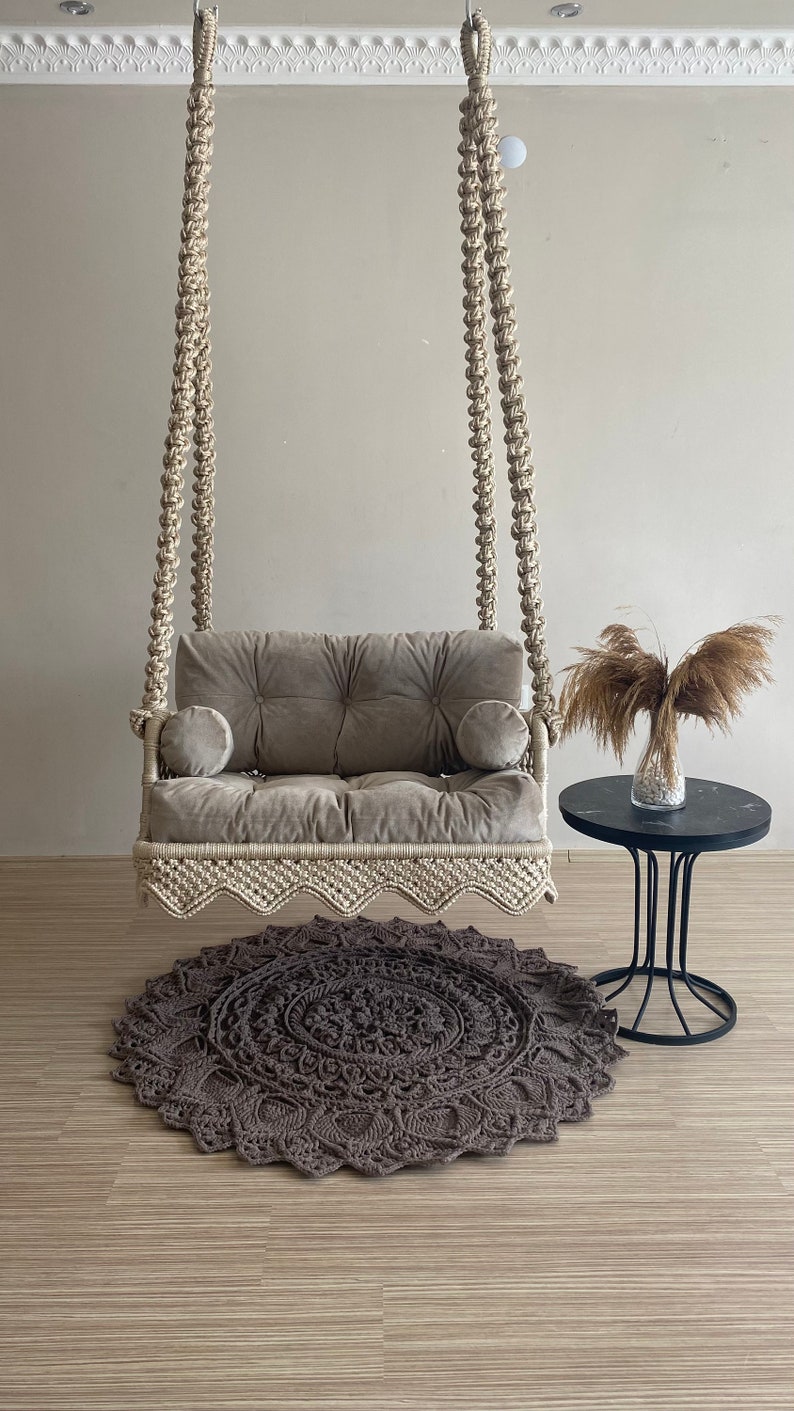 May include: A macrame swing chair with a gray cushion and two pillows. The swing is hanging from the ceiling and is over a brown rug with a circular pattern.